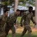 Three U.S. Army soldiers in MultiCam/OCP camouflage uniforms are running at speed in an outdoor wooded area. They wear shoulder patches with the number '2', indicating assignment to the 2nd Infantry Division or a similar unit. The image uses motion blur technique to convey speed and urgency.
