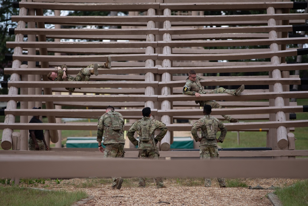 Military personnel in camouflage uniforms are navigating a large wooden obstacle course structure consisting of horizontal log beams arranged in a grid pattern. Some soldiers are actively climbing or traversing the upper levels of the structure, while others stand at the base observing or waiting their turn. This appears to be a military physical training or obstacle course exercise.
