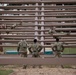 Military personnel in camouflage uniforms are navigating a large wooden obstacle course structure consisting of horizontal log beams arranged in a grid pattern. Some soldiers are actively climbing or traversing the upper levels of the structure, while others stand at the base observing or waiting their turn. This appears to be a military physical training or obstacle course exercise.