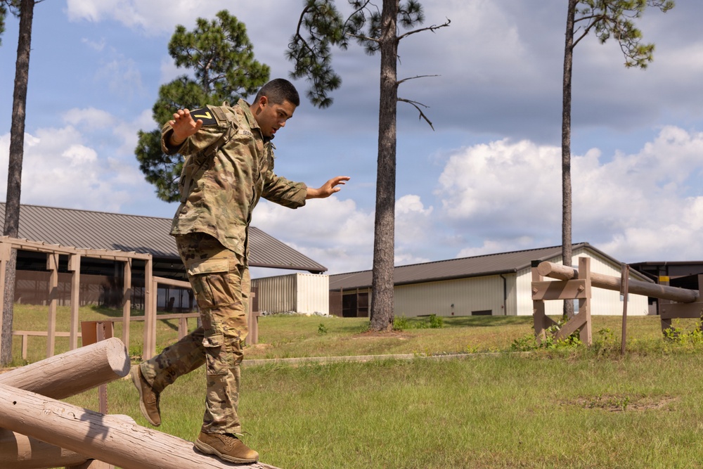 A soldier in U.S. Army OCP (Operational Camouflage Pattern) uniform is navigating a log obstacle course, balancing while stepping over or across wooden logs. The soldier wears a numbered bib (number 7), suggesting a timed competition or evaluation event. The setting appears to be a military installation obstacle course with barracks-style buildings visible in the background.