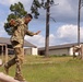 A soldier in U.S. Army OCP (Operational Camouflage Pattern) uniform is navigating a log obstacle course, balancing while stepping over or across wooden logs. The soldier wears a numbered bib (number 7), suggesting a timed competition or evaluation event. The setting appears to be a military installation obstacle course with barracks-style buildings visible in the background.