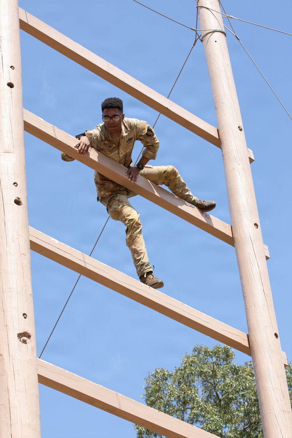 A soldier in U.S. Army camouflage (OCP/MultiCam) uniform is climbing a tall wooden obstacle course structure against a clear blue sky. The individual, wearing glasses and a name tape reading 'LANE', is mid-climb on a large wooden ladder-like frame supported by two vertical poles. This appears to be a military training exercise, likely at a basic training or ROTC obstacle course.