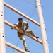 A soldier in U.S. Army camouflage (OCP/MultiCam) uniform is climbing a tall wooden obstacle course structure against a clear blue sky. The individual, wearing glasses and a name tape reading 'LANE', is mid-climb on a large wooden ladder-like frame supported by two vertical poles. This appears to be a military training exercise, likely at a basic training or ROTC obstacle course.