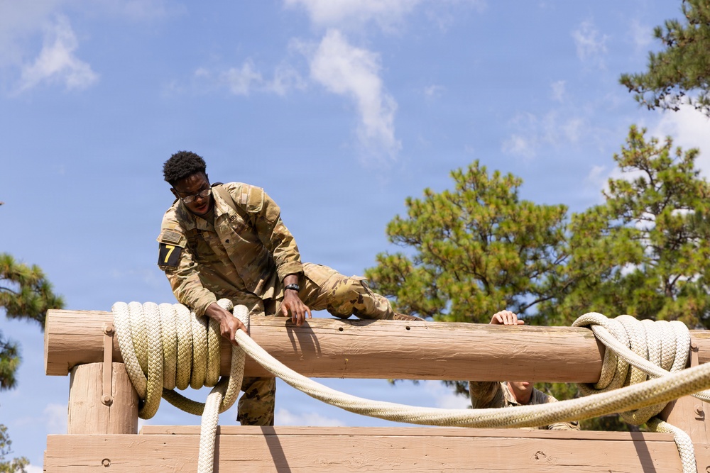 A soldier in camouflage uniform is navigating an elevated obstacle course element, climbing over a large horizontal log secured with thick rope. The individual wears a number '7' bib on their arm, suggesting a competitive or evaluated training event. Pine trees and a partly cloudy sky are visible in the background.
