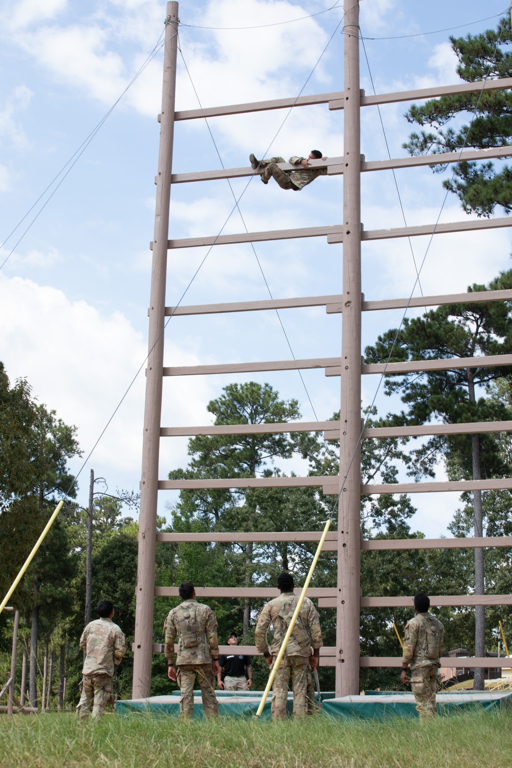 Military personnel are conducting obstacle course training at what appears to be a military installation. One soldier is near the top of a tall wooden ladder/climbing structure, while four others in camouflage uniforms observe from the ground below. The setting includes tall pine trees, safety mats at the base, and stabilizing cables attached to the structure.
