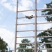 Military personnel are conducting obstacle course training at what appears to be a military installation. One soldier is near the top of a tall wooden ladder/climbing structure, while four others in camouflage uniforms observe from the ground below. The setting includes tall pine trees, safety mats at the base, and stabilizing cables attached to the structure.
