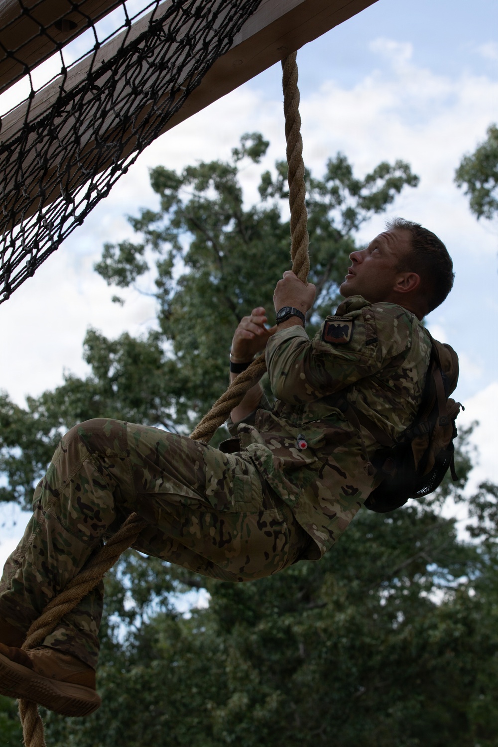 A soldier in MultiCam/OCP camouflage uniform is climbing a thick rope on a military obstacle course. The individual is looking upward while gripping the rope with both hands, with legs wrapped around it for support. A cargo net and wooden overhead structure are visible in the upper left, suggesting a formal military training facility.
