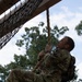 A soldier in MultiCam/OCP camouflage uniform is climbing a thick rope on a military obstacle course. The individual is looking upward while gripping the rope with both hands, with legs wrapped around it for support. A cargo net and wooden overhead structure are visible in the upper left, suggesting a formal military training facility.