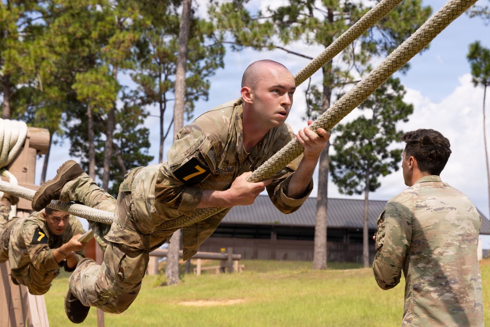 Military personnel in camouflage uniforms are navigating a rope obstacle course at what appears to be a military training facility. The primary subject, wearing a number '7' armband, is suspended horizontally while gripping a thick rope, demonstrating significant upper body strength and technique. An observer in matching uniform watches from the right side while another trainee performs the same obstacle in the background.