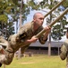 Military personnel in camouflage uniforms are navigating a rope obstacle course at what appears to be a military training facility. The primary subject, wearing a number '7' armband, is suspended horizontally while gripping a thick rope, demonstrating significant upper body strength and technique. An observer in matching uniform watches from the right side while another trainee performs the same obstacle in the background.