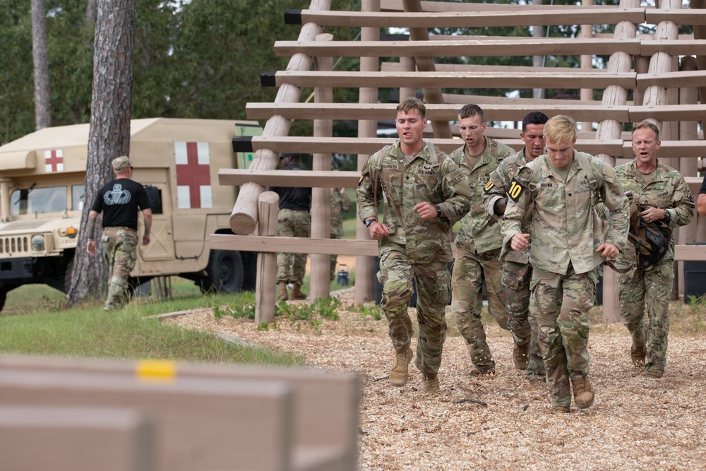 A group of U.S. Army soldiers in MultiCam/OCP camouflage uniforms are assisting a fellow soldier wearing a numbered bib (number 10), suggesting a military competition or assessment event. Several soldiers are physically supporting the numbered competitor as they move along a wood-chip path near a log obstacle course structure. A medical HMMWV (Humvee) with a Red Cross marking is visible in the background, indicating medical support is on standby.
