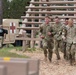 A group of U.S. Army soldiers in MultiCam/OCP camouflage uniforms are assisting a fellow soldier wearing a numbered bib (number 10), suggesting a military competition or assessment event. Several soldiers are physically supporting the numbered competitor as they move along a wood-chip path near a log obstacle course structure. A medical HMMWV (Humvee) with a Red Cross marking is visible in the background, indicating medical support is on standby.