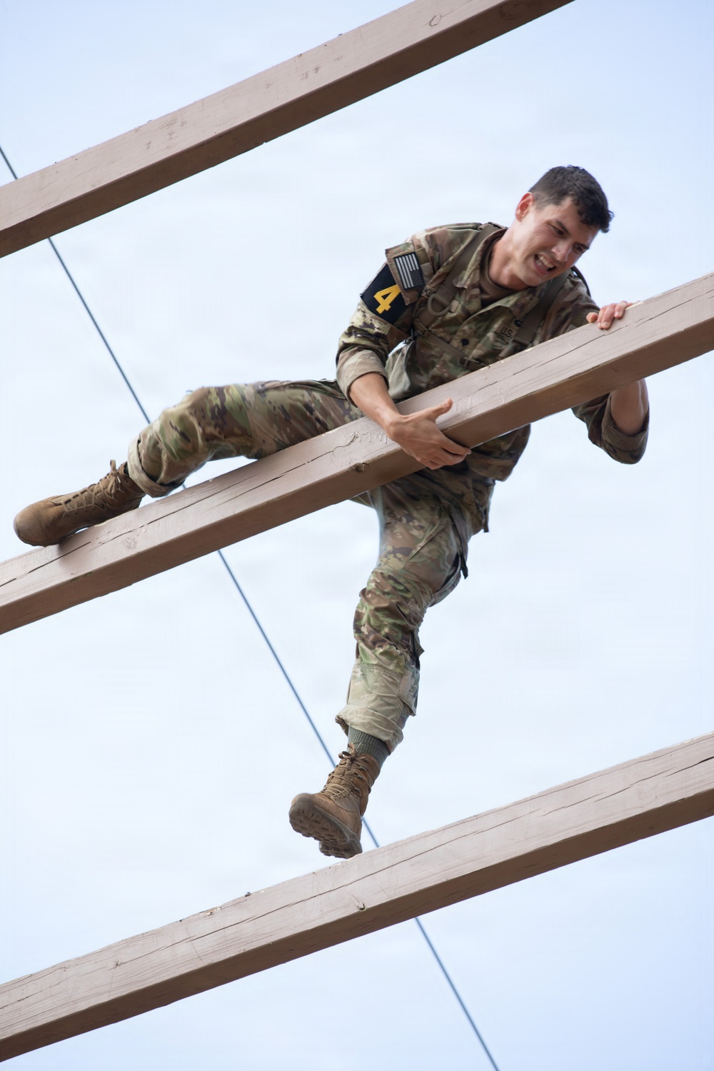 A soldier in US Army OCP (Operational Camouflage Pattern) uniform is navigating a military obstacle course, climbing over horizontal wooden beams elevated above the ground. The photo is taken from a low angle looking upward, capturing the soldier mid-movement with visible effort and a determined expression. A number '4' patch is visible on the shoulder, suggesting a competition or training event.