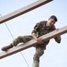 A soldier in US Army OCP (Operational Camouflage Pattern) uniform is navigating a military obstacle course, climbing over horizontal wooden beams elevated above the ground. The photo is taken from a low angle looking upward, capturing the soldier mid-movement with visible effort and a determined expression. A number '4' patch is visible on the shoulder, suggesting a competition or training event.