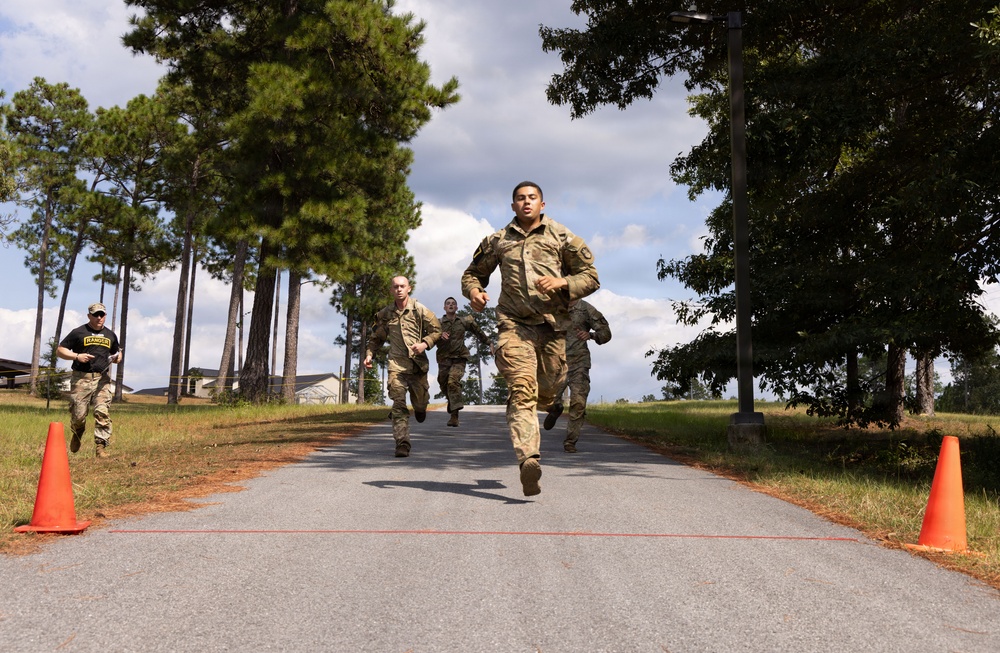 A group of military personnel in camouflage uniforms are running toward a finish line marked by a red cord stretched between two orange traffic cones. One individual in a black shirt with 'RANGER' text visible is observing or officiating on the left side. The setting appears to be a paved road lined with pine and deciduous trees on a military installation.