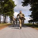 A group of military personnel in camouflage uniforms are running toward a finish line marked by a red cord stretched between two orange traffic cones. One individual in a black shirt with 'RANGER' text visible is observing or officiating on the left side. The setting appears to be a paved road lined with pine and deciduous trees on a military installation.