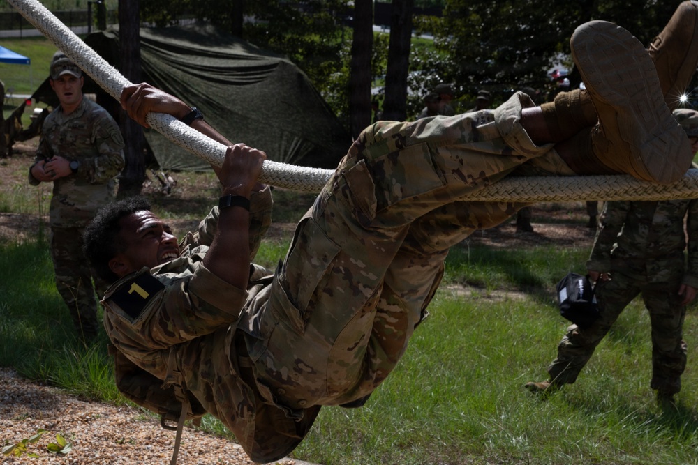A soldier in OCP (Operational Camouflage Pattern) uniform, marked with the number '1', is performing a rope traverse or obstacle course exercise, hanging horizontally from a thick rope. Several other soldiers in matching uniforms observe in the background near military tents. The setting appears to be an outdoor military training area with trees and grass.
