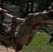 A soldier in OCP (Operational Camouflage Pattern) uniform, marked with the number '1', is performing a rope traverse or obstacle course exercise, hanging horizontally from a thick rope. Several other soldiers in matching uniforms observe in the background near military tents. The setting appears to be an outdoor military training area with trees and grass.