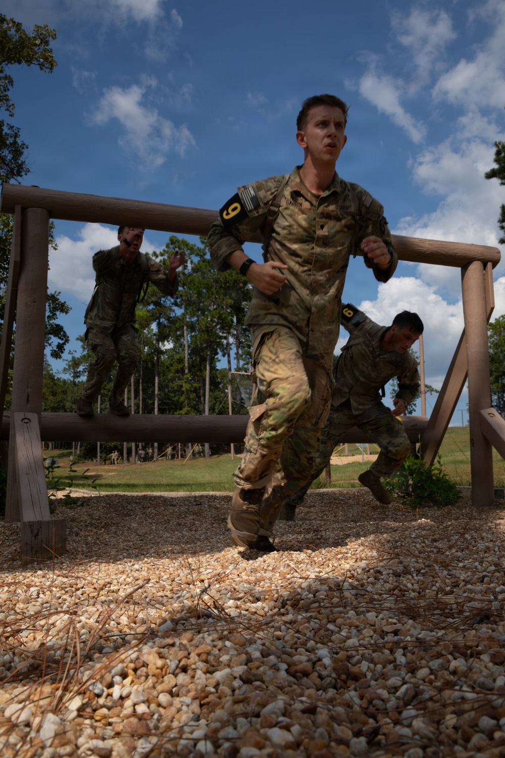 Military personnel in camouflage uniforms are navigating an outdoor obstacle course, ducking under or running past a horizontal log barrier. The foreground soldier wears a black armband with the number '9' and a name tape reading 'DARREN' along with a 'STUDENT' tape, suggesting a training exercise. At least three soldiers are visible at various stages of completing the obstacle.