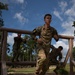 Military personnel in camouflage uniforms are navigating an outdoor obstacle course, ducking under or running past a horizontal log barrier. The foreground soldier wears a black armband with the number '9' and a name tape reading 'DARREN' along with a 'STUDENT' tape, suggesting a training exercise. At least three soldiers are visible at various stages of completing the obstacle.