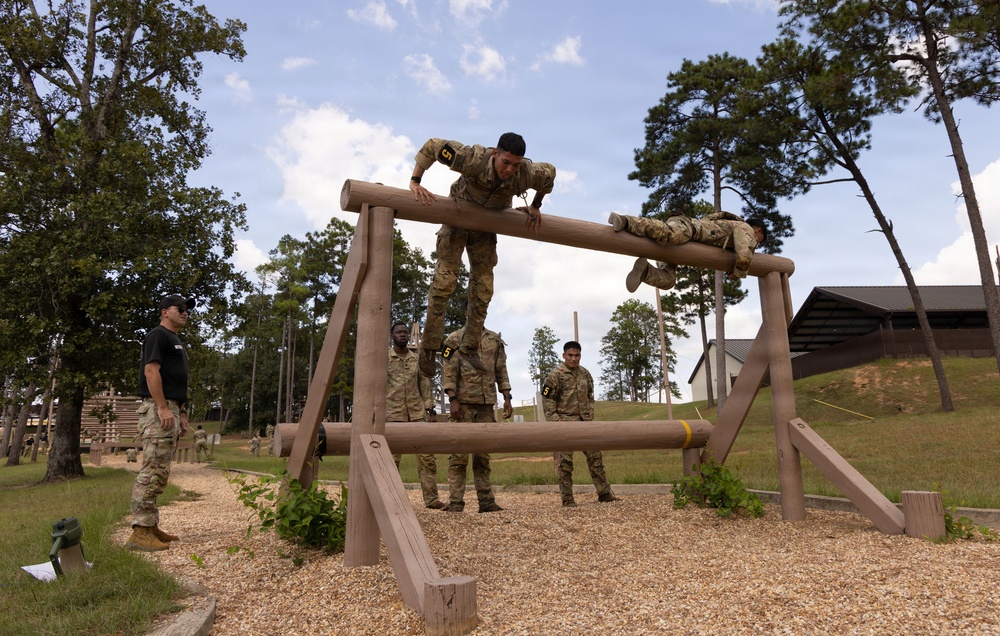 Military personnel are navigating a wooden obstacle course at what appears to be a military training facility. Two soldiers are actively on the horizontal log obstacle — one climbing over the top and one crawling across — while several others stand nearby observing or waiting their turn. An instructor in a black shirt monitors the activity from the left side.