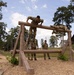 Military personnel are navigating a wooden obstacle course at what appears to be a military training facility. Two soldiers are actively on the horizontal log obstacle — one climbing over the top and one crawling across — while several others stand nearby observing or waiting their turn. An instructor in a black shirt monitors the activity from the left side.