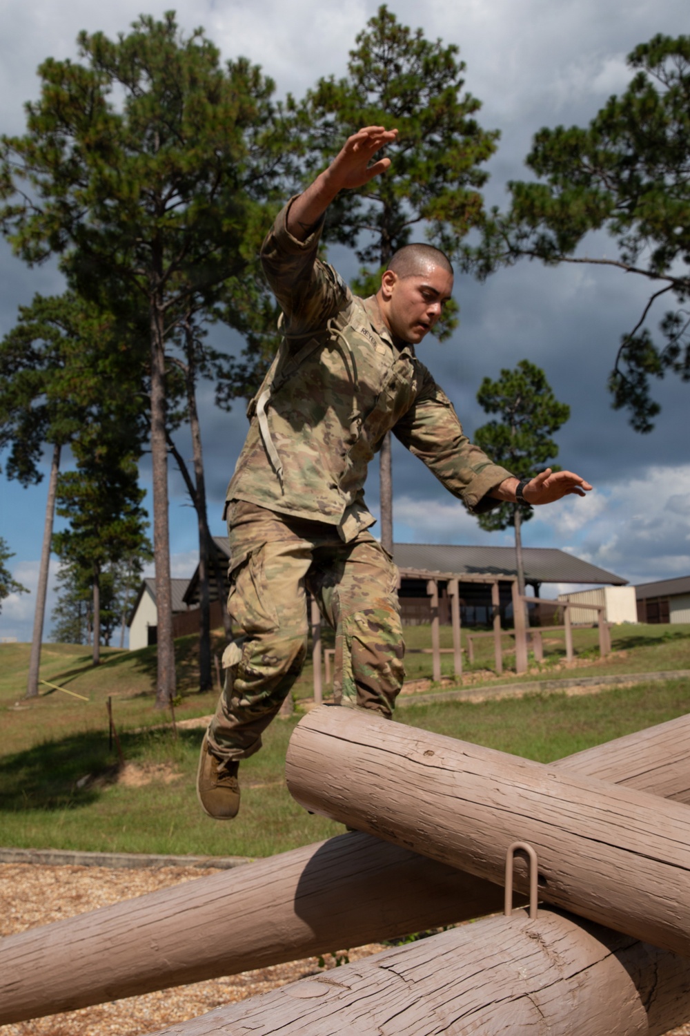 A soldier in US Army OCP (Operational Camouflage Pattern) uniform is navigating a military obstacle course, specifically jumping or balancing over crossed log obstacles. The individual appears to be mid-movement with arms extended for balance, suggesting active physical training. The setting is an outdoor military training facility with buildings and pine trees visible in the background.