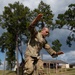 A soldier in US Army OCP (Operational Camouflage Pattern) uniform is navigating a military obstacle course, specifically jumping or balancing over crossed log obstacles. The individual appears to be mid-movement with arms extended for balance, suggesting active physical training. The setting is an outdoor military training facility with buildings and pine trees visible in the background.