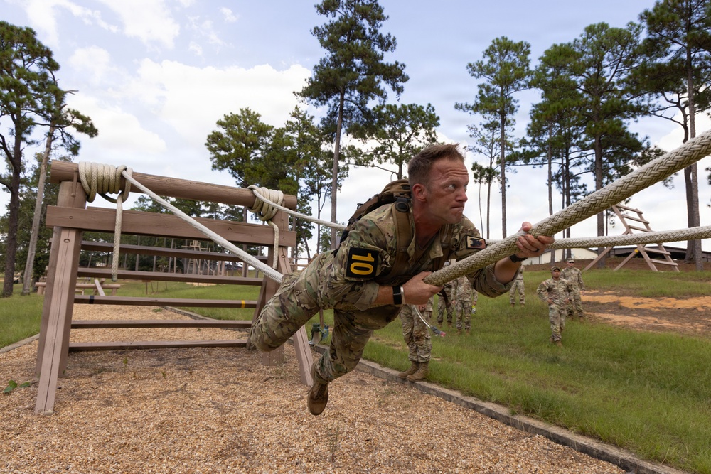 A soldier wearing camouflage uniform with the number '10' on their back is traversing a rope obstacle on a military training course. The individual is suspended horizontally, gripping a thick braided rope with both hands while their body is nearly parallel to the ground. Several other uniformed personnel are visible in the background observing or waiting their turn.