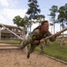 A soldier wearing camouflage uniform with the number '10' on their back is traversing a rope obstacle on a military training course. The individual is suspended horizontally, gripping a thick braided rope with both hands while their body is nearly parallel to the ground. Several other uniformed personnel are visible in the background observing or waiting their turn.