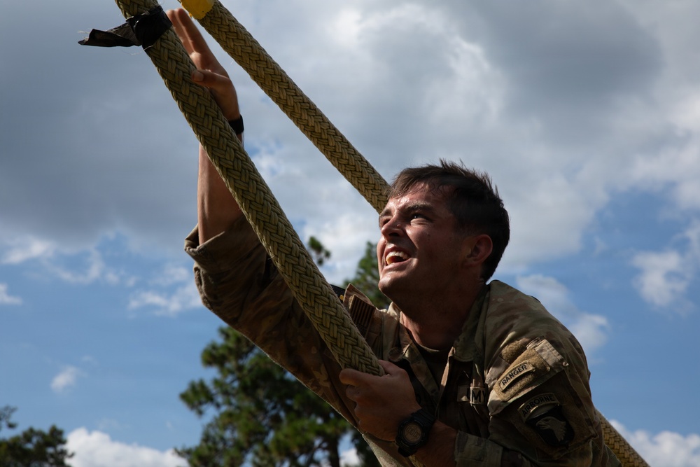 A U.S. Army soldier is climbing a thick braided rope during what appears to be a military training exercise or obstacle course. The soldier is wearing a multicam uniform with visible patches including a 'RANGER' tab and what appears to be a 101st Airborne Division patch. The soldier's expression shows intense physical exertion with a grimacing smile.