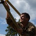A U.S. Army soldier is climbing a thick braided rope during what appears to be a military training exercise or obstacle course. The soldier is wearing a multicam uniform with visible patches including a 'RANGER' tab and what appears to be a 101st Airborne Division patch. The soldier's expression shows intense physical exertion with a grimacing smile.