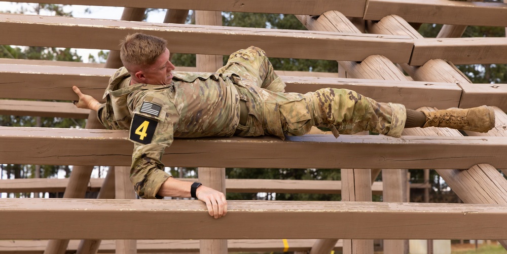 A U.S. Army soldier wearing OCP (Operational Camouflage Pattern) uniform and numbered bib '4' navigates a military obstacle course, crawling horizontally across wooden beam structures. The soldier appears to be low-crawling or maneuvering between parallel wooden beams, likely part of a timed competition or training evaluation. Trees and an overcast sky are visible in the background.