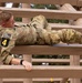 A U.S. Army soldier wearing OCP (Operational Camouflage Pattern) uniform and numbered bib '4' navigates a military obstacle course, crawling horizontally across wooden beam structures. The soldier appears to be low-crawling or maneuvering between parallel wooden beams, likely part of a timed competition or training evaluation. Trees and an overcast sky are visible in the background.
