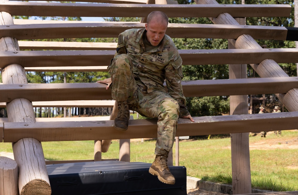 A U.S. Army soldier in OCP (Operational Camouflage Pattern) uniform is navigating a wooden obstacle course structure, appearing to climb through or over horizontal wooden beams supported by log posts. The soldier is actively engaged in a physical fitness or training exercise, likely at a military installation obstacle course. The setting is an outdoor training area with trees and grass visible in the background.