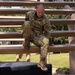A U.S. Army soldier in OCP (Operational Camouflage Pattern) uniform is navigating a wooden obstacle course structure, appearing to climb through or over horizontal wooden beams supported by log posts. The soldier is actively engaged in a physical fitness or training exercise, likely at a military installation obstacle course. The setting is an outdoor training area with trees and grass visible in the background.