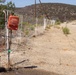 The image shows a rural electric fence installation along a dry, arid dirt road in what appears to be a semi-desert or chaparral environment. A red Speedrite 3000 electric fence energizer is mounted on a white fence post, connected to a battery unit on the ground. The landscape features dry scrubland, a hillside in the background, and a utility pole visible along the road.