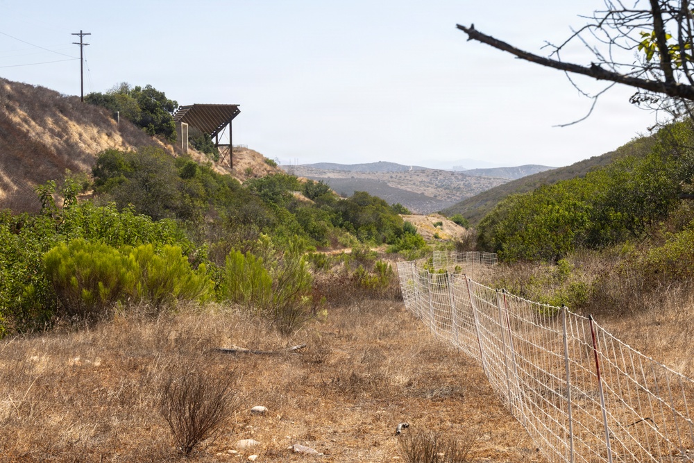 A dry, semi-arid hillside landscape featuring a wire mesh fence running diagonally through the foreground into the middle distance. A wooden pergola or shade structure sits atop a hill to the left, alongside a utility pole with power lines. The terrain is characteristic of Southern California chaparral, with dry grasses, scrub brush, and rolling hills visible in the background.