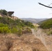 A dry, semi-arid hillside landscape featuring a wire mesh fence running diagonally through the foreground into the middle distance. A wooden pergola or shade structure sits atop a hill to the left, alongside a utility pole with power lines. The terrain is characteristic of Southern California chaparral, with dry grasses, scrub brush, and rolling hills visible in the background.