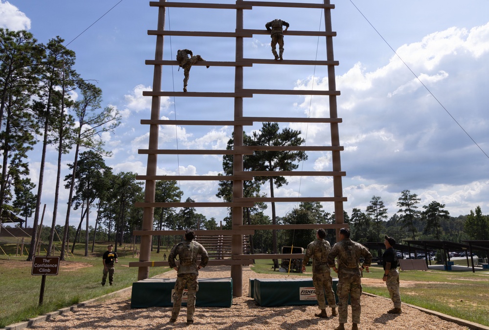 Military personnel are training on a tall wooden obstacle called 'The Confidence Climb,' a large vertical ladder structure used in military training courses. Two soldiers are actively climbing near the top of the structure while several others observe from the ground below. The setting appears to be a military training facility surrounded by pine trees under a partly cloudy sky.