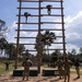 Military personnel are training on a tall wooden obstacle called 'The Confidence Climb,' a large vertical ladder structure used in military training courses. Two soldiers are actively climbing near the top of the structure while several others observe from the ground below. The setting appears to be a military training facility surrounded by pine trees under a partly cloudy sky.