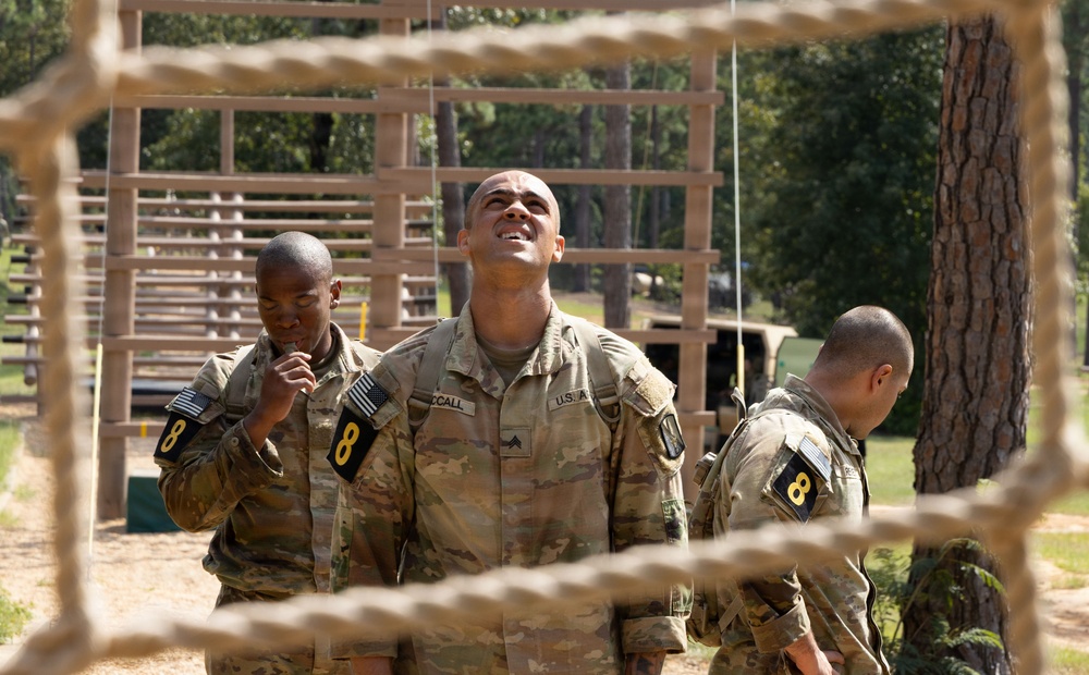 Three U.S. Army soldiers in OCP (Operational Camouflage Pattern) uniforms are photographed through a rope net obstacle at what appears to be a military training course. All three wear the number '8' patch on their left shoulders, suggesting they belong to the same unit or group. The central soldier, identified by nametape as 'McCall,' is looking upward, possibly assessing an overhead obstacle.