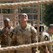 Three U.S. Army soldiers in OCP (Operational Camouflage Pattern) uniforms are photographed through a rope net obstacle at what appears to be a military training course. All three wear the number '8' patch on their left shoulders, suggesting they belong to the same unit or group. The central soldier, identified by nametape as 'McCall,' is looking upward, possibly assessing an overhead obstacle.