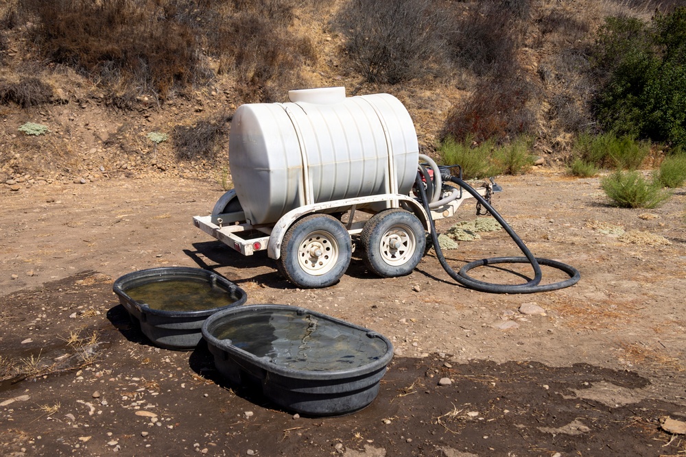 A white plastic water tank mounted on a dual-axle trailer is positioned on dry, rocky terrain in an arid landscape. A black hose connects the tank to two dark rubber livestock water troughs placed on the ground in front of the trailer. The setting suggests a water delivery operation for livestock or wildlife in a remote, drought-affected area.
