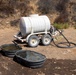 A white plastic water tank mounted on a dual-axle trailer is positioned on dry, rocky terrain in an arid landscape. A black hose connects the tank to two dark rubber livestock water troughs placed on the ground in front of the trailer. The setting suggests a water delivery operation for livestock or wildlife in a remote, drought-affected area.