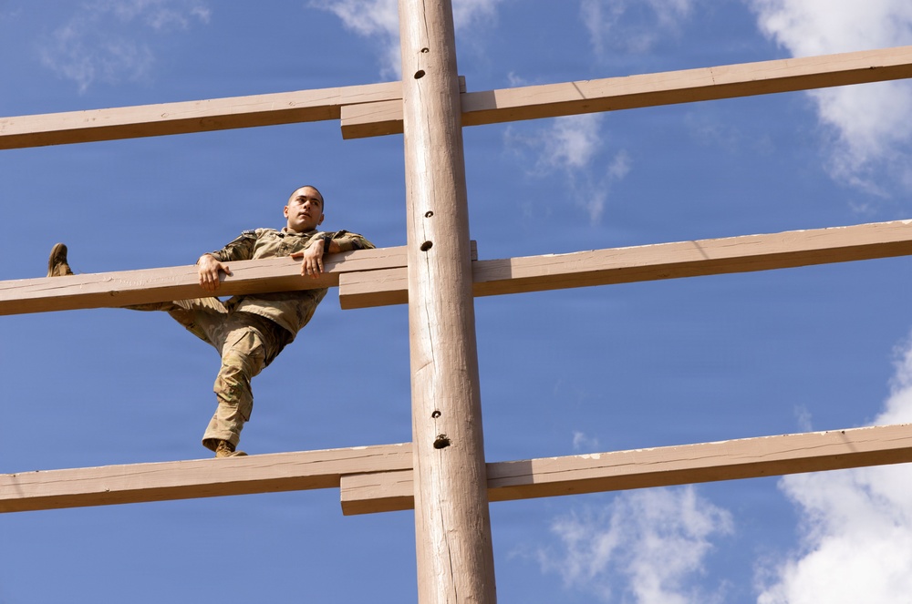 A soldier in camouflage uniform is climbing a large wooden obstacle structure, photographed from a low angle looking upward. The structure consists of horizontal wooden planks attached to a central vertical pole, resembling a military training obstacle course element. The blue sky with scattered clouds serves as the background.