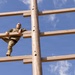 A soldier in camouflage uniform is climbing a large wooden obstacle structure, photographed from a low angle looking upward. The structure consists of horizontal wooden planks attached to a central vertical pole, resembling a military training obstacle course element. The blue sky with scattered clouds serves as the background.