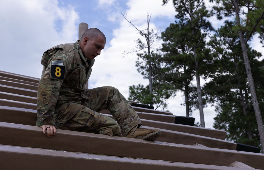 A soldier in U.S. Army OCP (Operational Camouflage Pattern) uniform is navigating what appears to be a wooden obstacle course structure, climbing or traversing angled wooden beams. The soldier wears a number '8' patch on their left sleeve along with a U.S. flag patch, suggesting a competitive or training event. The setting is an outdoor military training environment surrounded by pine trees.