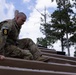 A soldier in U.S. Army OCP (Operational Camouflage Pattern) uniform is navigating what appears to be a wooden obstacle course structure, climbing or traversing angled wooden beams. The soldier wears a number '8' patch on their left sleeve along with a U.S. flag patch, suggesting a competitive or training event. The setting is an outdoor military training environment surrounded by pine trees.