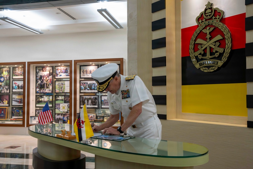 A senior military officer in white dress uniform is signing a document or guest book at a reception desk in what appears to be a Brunei Armed Forces headquarters or official military facility. The setting features the Royal Brunei Armed Forces emblem and flag prominently displayed on the wall, alongside small US and Brunei flags on the desk, indicating a bilateral military visit or diplomatic engagement. The background displays framed photographs suggesting a history gallery or ceremonial reception area.